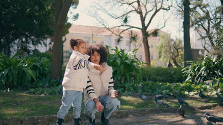 Little girl feeding birds in green sunny park with smiling mom. Cute african american daughter with mother throwing seeds to hungry pigeons. Flock gray doves flying overhead carefree happy family.の写真素材