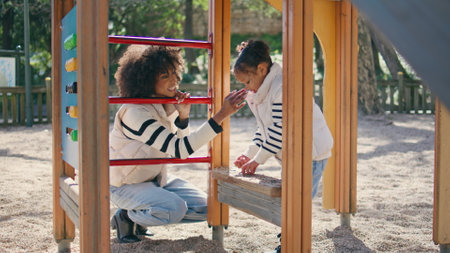 Woman looking daughter game standing on sunny playground. Cheerful african american mother watching...