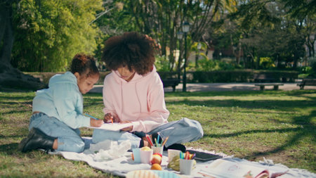 Creative family drawing on picnic at beautiful green lawn sunny weekend. Relaxed african american mother painting on paper with cute little daughter enjoying art. Joyful mom child creating picture.の写真素材
