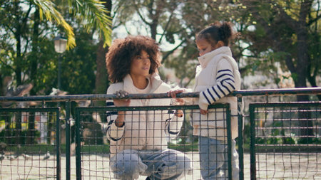 Family looking on flying pigeons standing at park fence sunny day. Smiling african american woman enjoying carefree weekend with cute little daughter outdoors. Happy curly girl watching wild birdsの写真素材