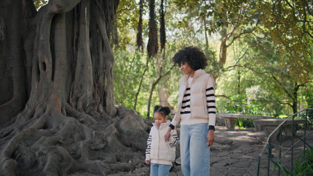 Woman daughter strolling park near old beautiful tree sunny day. Relaxed african american mother holding girl hand walking on path. Cute mom child wearing identical stylish look enjoy weekend togetherの写真素材
