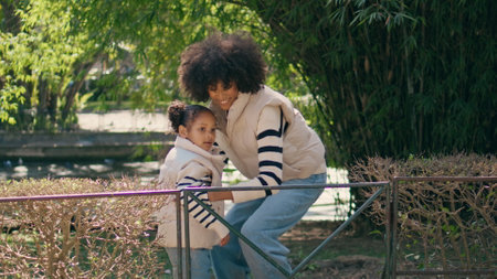 Woman carrying daughter on walk transplanting over fence. Cheerful african american mother taking little girl on hands strolling at sunny park together. Happy mom have fun on nature with cute child.の写真素材