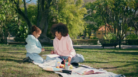 Family sitting picnic blanket on sunny green nature having fun together. Cute african american child drawing on paper with happy curly mother. Relaxed little girl enjoying creativity on meadow grass.の写真素材