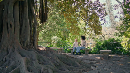 Unrecognizable mother daughter sitting on park bench looking at beautiful sprawling tree. Happy relaxed woman enjoy weekend rest with cute kid in green forest. Happy african american family relaxing.の写真素材