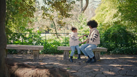 Mother clapping hands with daughter sitting bench beautiful green park. Young african american mom play funny games with cute little girl on sunny nature. Carefree family enjoy time happy together.の写真素材