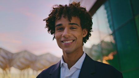 Smiling businessman posing sunset city portrait. Curly young man looking camera standing in evening business district closeup. Joyful latin entrepreneur face staring lens feeling happy on dusk streetの写真素材