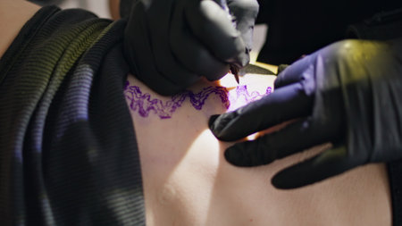 Master hands drawing tattoo at studio closeup. Unknown artist using pen tool wearing sterile black gloves at workplace. Person fingers working on unrecognizable client skin forming creative ink linesの写真素材