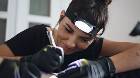 Smiling master transferring sketch at studio closeup. Positive tattoo artist holding pen in sterile gloves working on client skin. Cheerful head flashlight woman forming lines on unknown customer bodyの写真素材