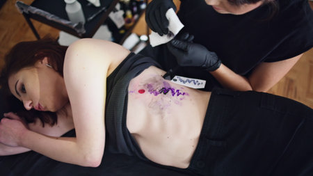 Skilful tattooer wiping client skin at studio closeup. Unrecognizable woman removing ink with tissue from customer body. Attentive tattoo artist creating black outline ending work at home salonの写真素材
