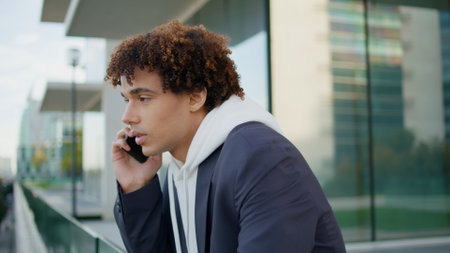 Serious teenager talking mobile at glass terrace closeup. Focused guy having phone call at modern architecture place. Young man speaking at building wall background. Curly hipster chatting smartphoneの写真素材