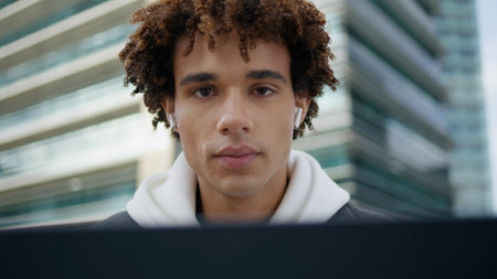 Earphones youngster using laptop computer at street close up. Curly teen looking camera attentively outdoor portrait. Handsome latin model holding gadget sitting at building background. People conceptの写真素材