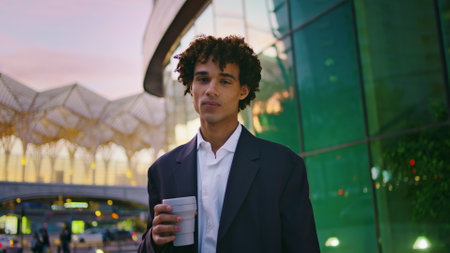 Happy man looking camera in business district closeup. Young businessman holding coffee posing at evening mall architecture. White collar guy enjoying hot beverage after work in urban setting portraitの写真素材