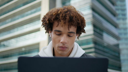 Focused guy working laptop outdoors portrait. Earphones student studying alone at architecture background closeup. Curly hair model using digital device in buildings downtown. Latin teenager learningの写真素材