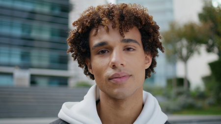 Young man looking camera in downtown closeup. Smiling latin guy staring lens on architecture background. Confident model face posing in urban district portrait. Contemporary teenager standing streetの写真素材