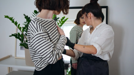 Back view client looking mirror at tattoo studio. Skilful master applying temporary picture on female body closeup. Serious tattooist and customer lady staring reflection preparing for procedureの写真素材
