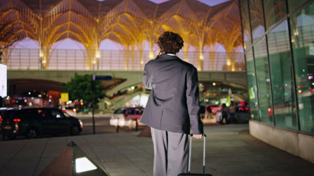 Businessman having phone call at night street. Unknown curly manager carrying baggage walking dark downtown back view. Formal clothes tourist arriving at new city. Guy going towards traffic roadの写真素材