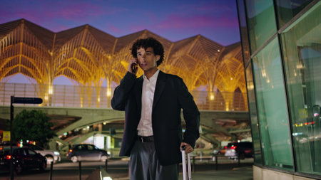 Young man talking phone at illuminated city. Smiling businessman calling smartphone waiting in airport bus stop. Curly entrepreneur waving arm meeting relatives in downtown district. Business tripの写真素材