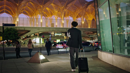 Suit man calling phone in dark street back view. Formal wear businessman carrying luggage walking at night road exterior. Business traveler pulling suitcase from airport terminal at urban backgroundの写真素材