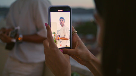 Woman hands recording video of man evening balcony. Closeup couple resting together at weekend. Girl silhouette listening ukulele music filming mobile. Relaxed guy play guitar at sunset terraceの写真素材