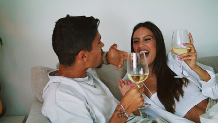 Laughing woman treating husband at sofa closeup. Overjoyed wife giving alcohol beverage to man at cozy place. Relaxed lovers flirting on romantic evening. Happy sweethearts having fun on vacationの写真素材