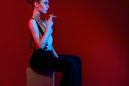 young sexy topless posing in ultra trendy fashionable pants with suspenders on a burgundy background with colored lightの写真素材