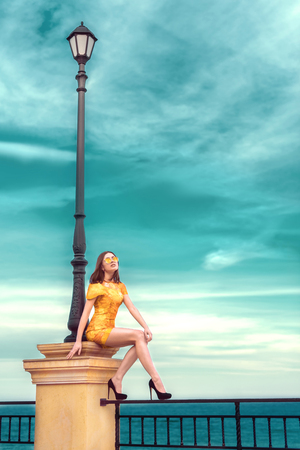 stylish and fashionable young brunette girl in yellow spring color dress posing on the beach against the seaの写真素材