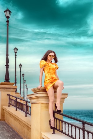 stylish and fashionable young brunette girl in yellow spring color dress posing on the beach against the seaの写真素材