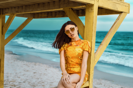 stylish and fashionable young brunette girl in yellow spring color dress posing on the beach against the seaの写真素材