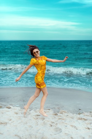 stylish and fashionable young brunette girl in yellow spring color dress posing on the beach against the seaの写真素材