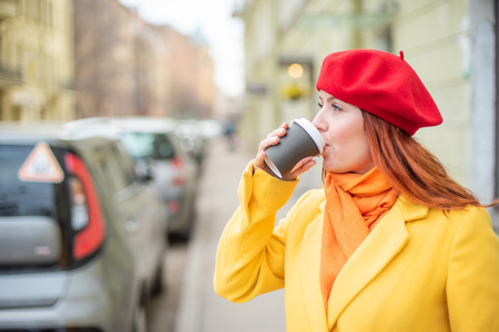 The red-haired young woman in a yellow coat and red beret is drinking coffee on the street to keep warmの写真素材