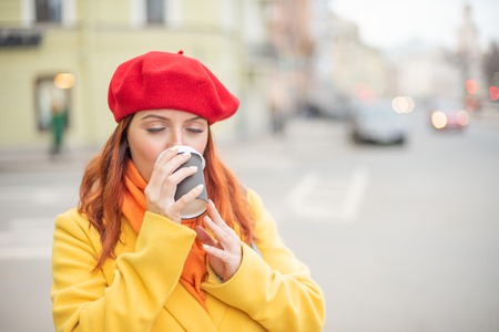 The red-haired young woman in a yellow coat and red beret is drinking coffee on the street to keep warmの写真素材