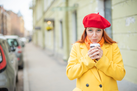 The red-haired young woman in a yellow coat and red beret is drinking coffee on the street to keep warmの写真素材