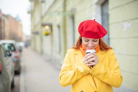 The red-haired young woman in a yellow coat and red beret is drinking coffee on the street to keep warmの写真素材
