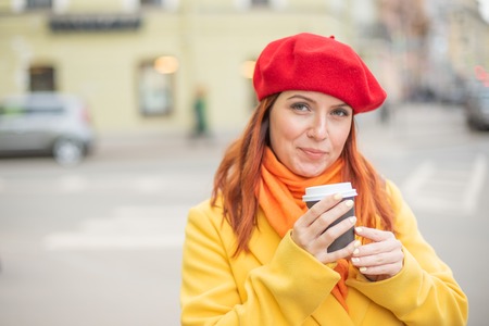 The red-haired young woman in a yellow coat and red beret is drinking coffee on the street to keep warmの写真素材