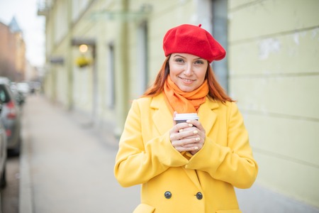 The red-haired young woman in a yellow coat and red beret is drinking coffee on the street to keep warmの写真素材