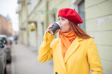The red-haired young woman in a yellow coat and red beret is drinking coffee on the street to keep warmの写真素材