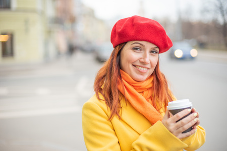 The red-haired young woman in a yellow coat and red beret is drinking coffee on the street to keep warmの写真素材