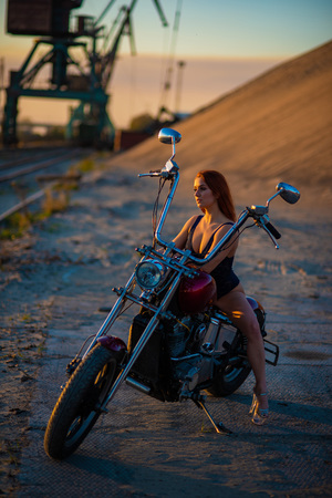 Red-haired woman in sexy lingerie in high heels sits on a motorcycle. Attractive red-haired girl sits on a motorcycle at sunset on the background of the industrial landscapeの写真素材
