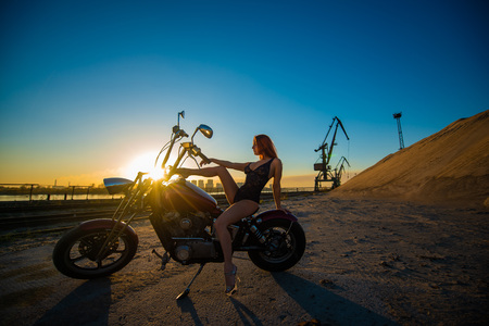 Red-haired woman in sexy lingerie in high heels sits on a motorcycle. Attractive red-haired girl sits on a motorcycle at sunset on the background of the industrial landscapeの写真素材
