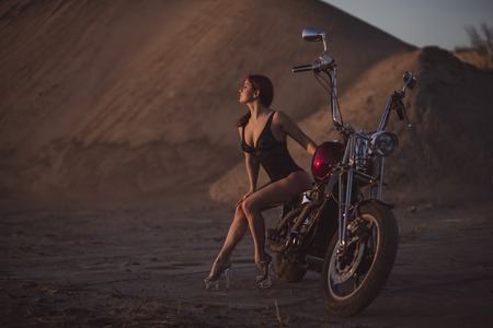Red-haired woman in sexy lingerie in high heels sits on a motorcycle. Attractive red-haired girl sits on a motorcycle at sunset on the background of the industrial landscapeの写真素材