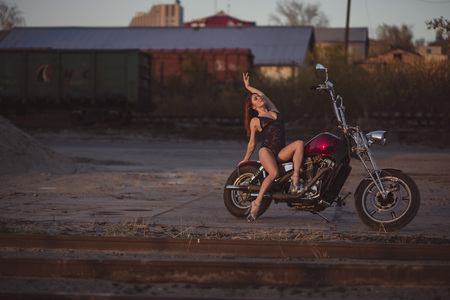Red-haired woman in sexy lingerie in high heels sits on a motorcycle. Attractive red-haired girl sits on a motorcycle at sunset on the background of the industrial landscapeの写真素材