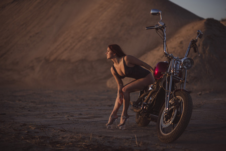 Red-haired woman in sexy lingerie in high heels sits on a motorcycle. Attractive red-haired girl sits on a motorcycle at sunset on the background of the industrial landscapeの写真素材