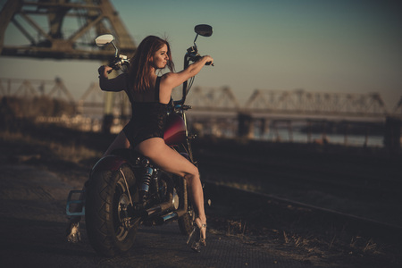 Red-haired woman in sexy lingerie in high heels sits on a motorcycle. Attractive red-haired girl sits on a motorcycle at sunset on the background of the industrial landscapeの写真素材