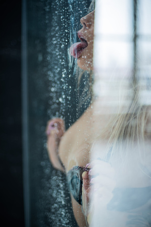European girl in black underwear, pressed her chest and hands to the wet glass and erotic licking tongue drops.の写真素材