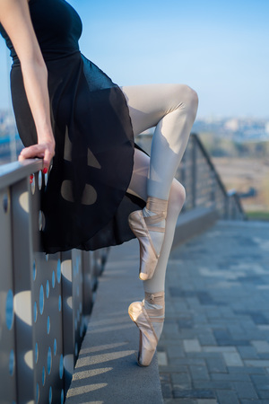 Ballerina in ballet legs in shoes and black tutu dancing by the fence. Beautiful young woman in black dress and pointe dancing outside. Gorgeous ballerina performing a dance outdoors. Close up.の写真素材