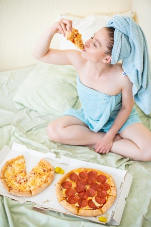 Cute girl with a towel on her head eats pizza in bed. Young woman eating fast food in bed. Bodypositive. Lots of italian food.の写真素材