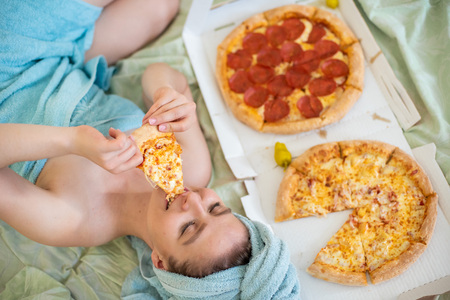 Cute girl with a towel on her head eats pizza in bed. Young woman eating pizza in bed. Life is a pleasure, body positive. Love for Italian food. Food habits, addiction to fast foodの写真素材
