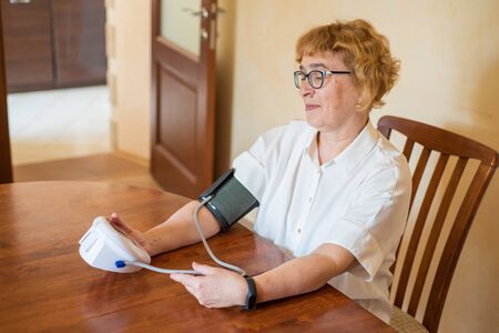 An elderly woman with glasses measures blood pressure using an electrical device in a bright room....