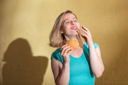 Portrait of a cute girl in a green dress walking outdoors and eating a burger on a warm summer day. Beautiful blonde is enjoying fast food at the yellow wall outside. Wrong food habitsの写真素材
