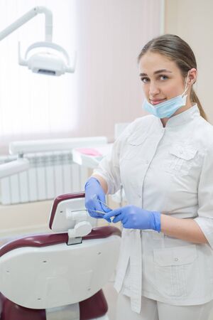 Attractive smiling female doctor in a white uniform at the workplace. Young beautiful female dentist in mask and gloves is standing in the office near the armchair with tools in her hands.の写真素材